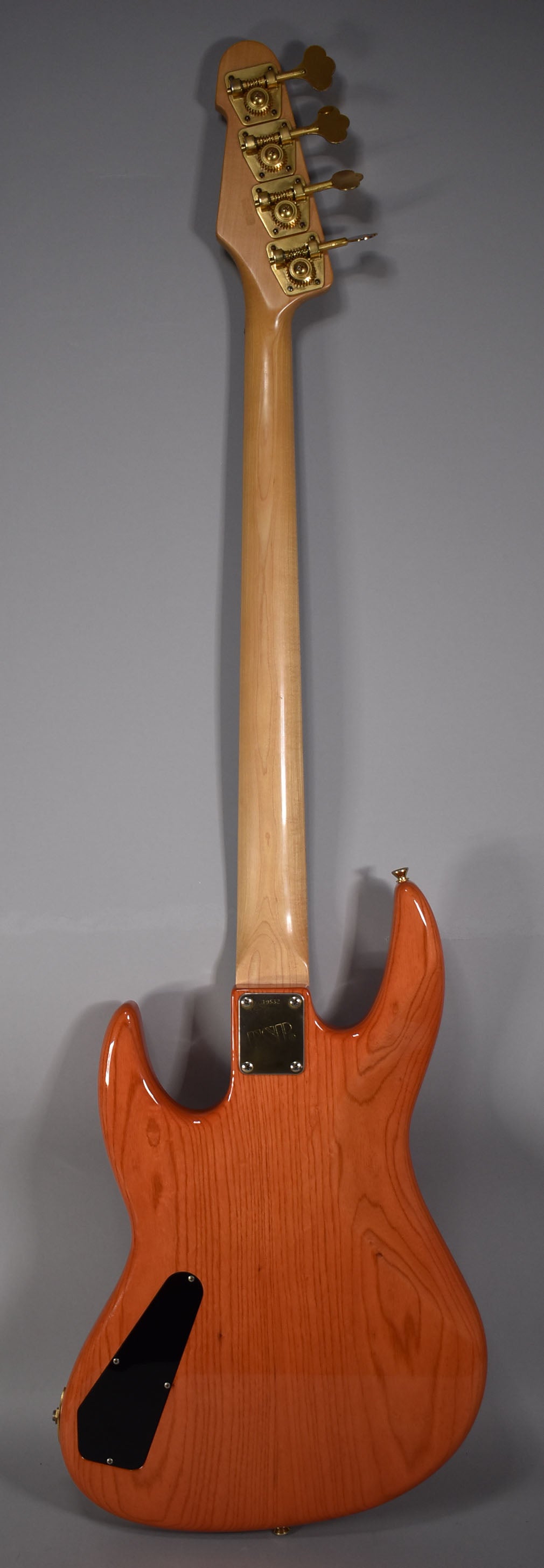 The back of a translucent orange bass with a light wood neck, gold tuners, and a black control cavity cover.