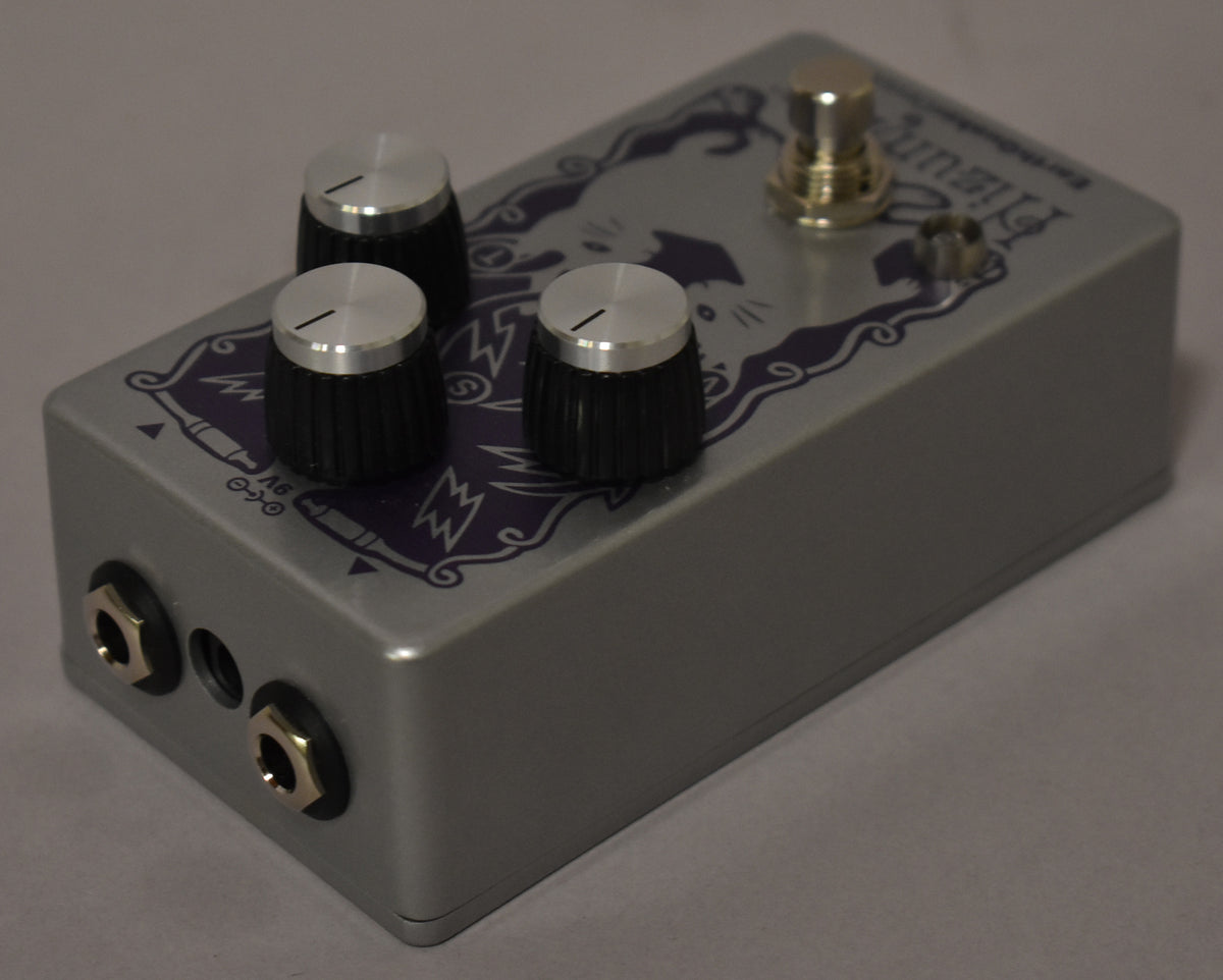 Silver pedal with purple cat graphic, three black knurled knobs with silver tops, a gold footswitch, and two input jacks.