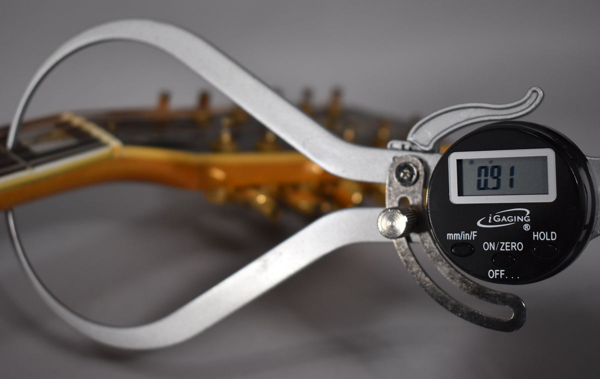 A digital caliper measures the guitar's nut width at 0.91, with the blonde headstock and gold tuners blurred in the background.