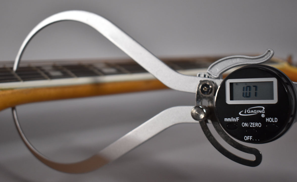 A digital caliper measures the neck relief of the natural blonde guitar, showing "1.07" on its display.