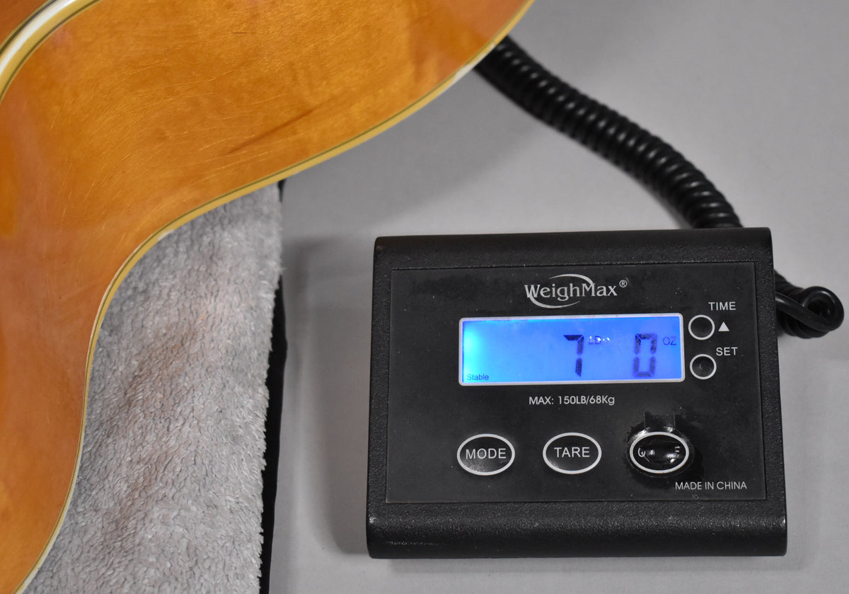 The guitar's blonde body, showing light wear, rests on a gray towel next to a digital scale displaying "7 lbs 0 oz".