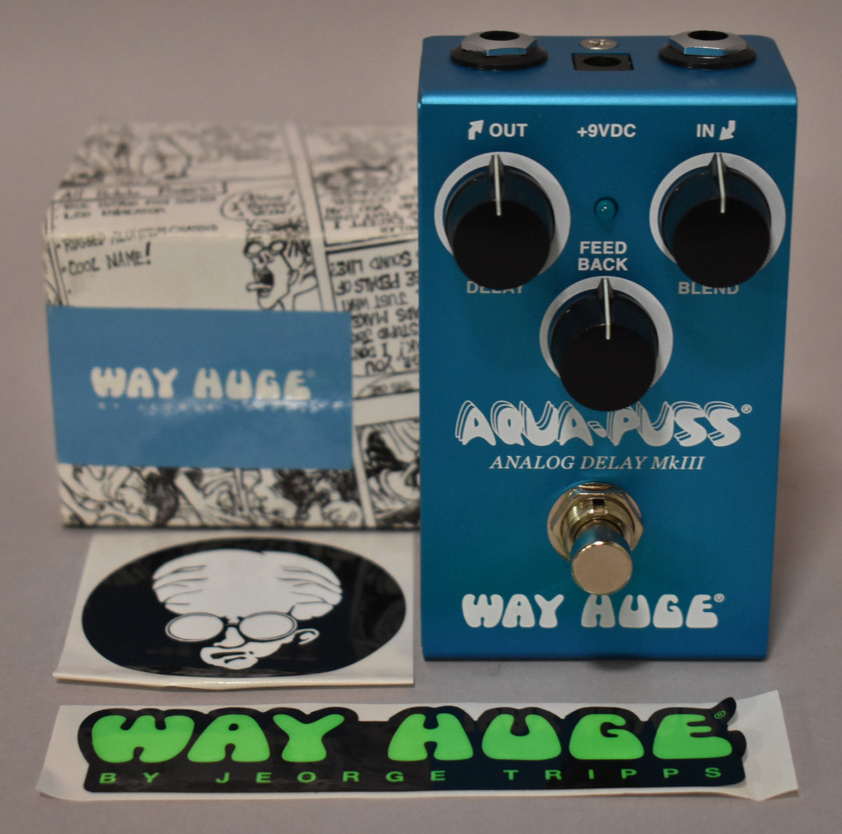 A teal pedal with three black knobs, a gold footswitch, and a comic-strip box with a blue band.