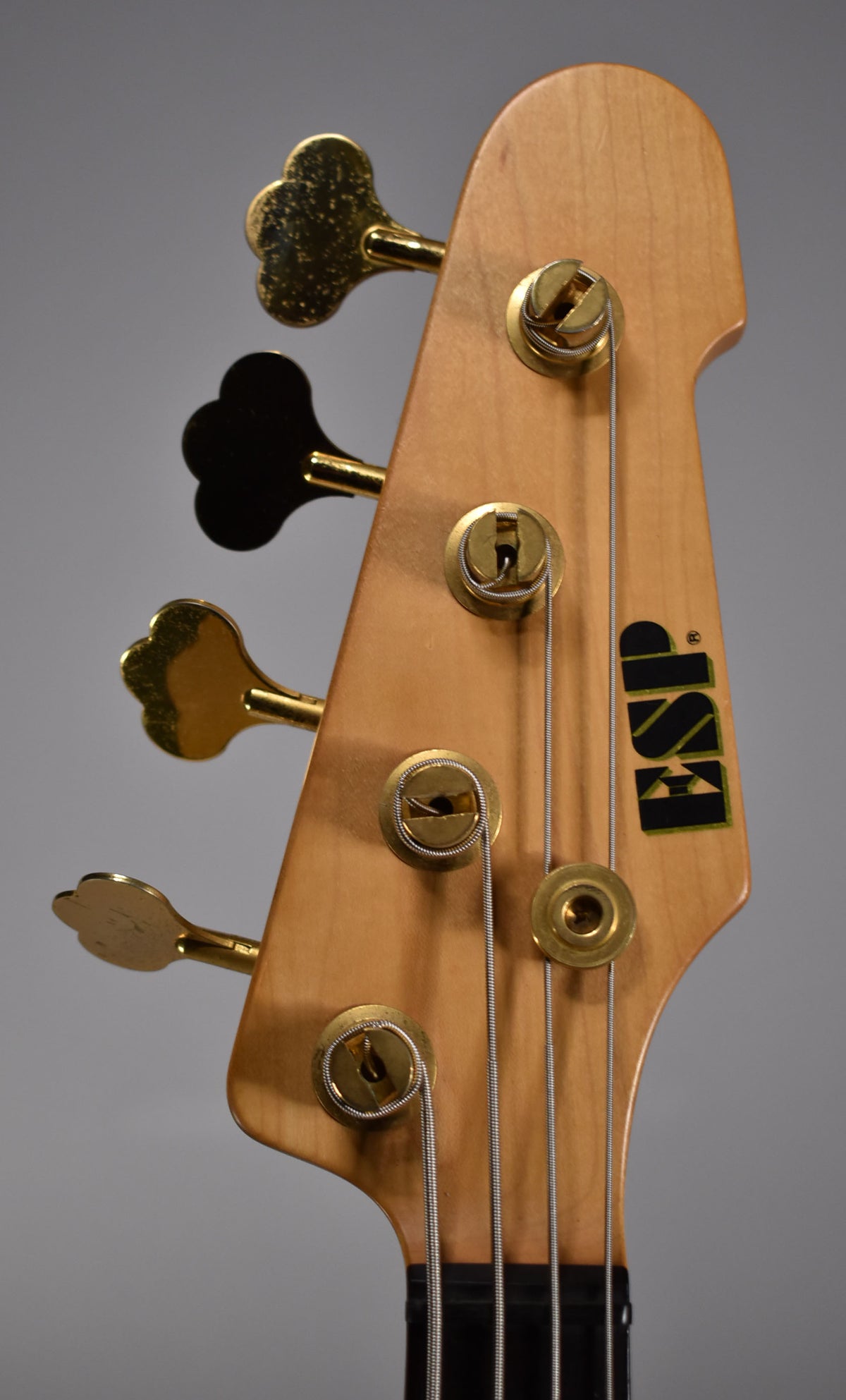 Close-up of the bass headstock, showing four gold clover-leaf tuners, three with visible wear, and a black logo.