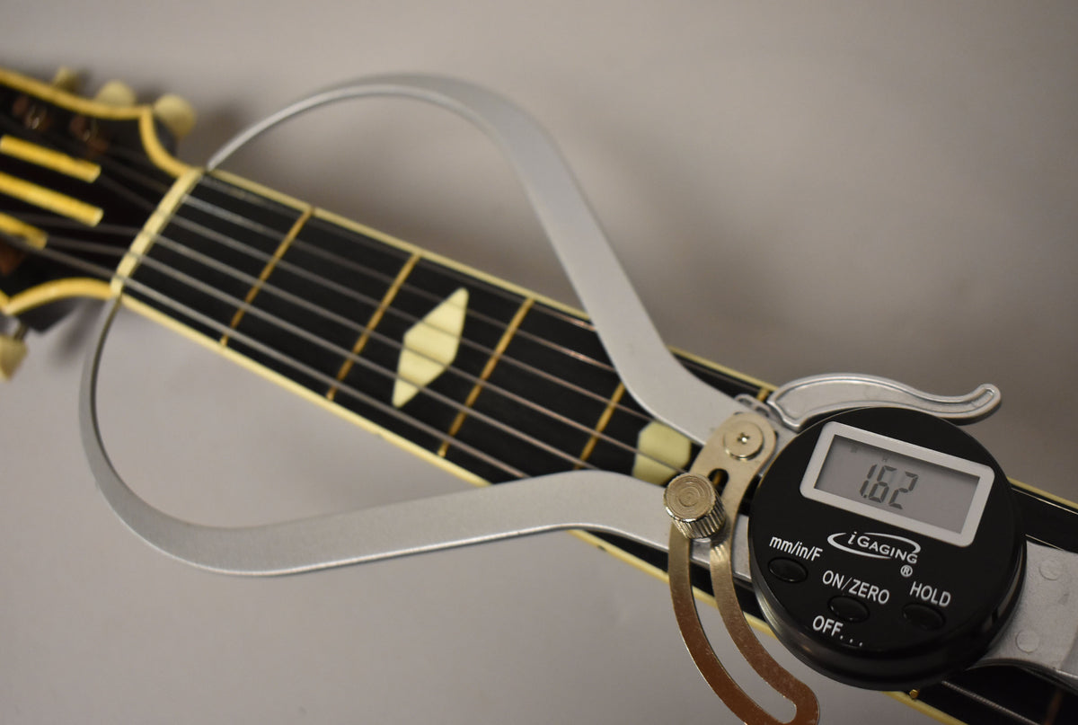 A close-up of the guitar's dark fretboard with diamond inlays, showing a digital caliper measuring the nut width at 1.62 inches.