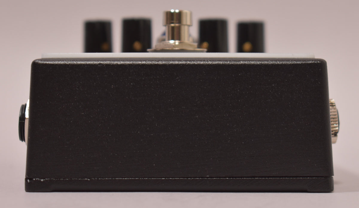 The black textured pedal is viewed from the side, showing the input and output jacks, with a few small chips along the bottom edge.