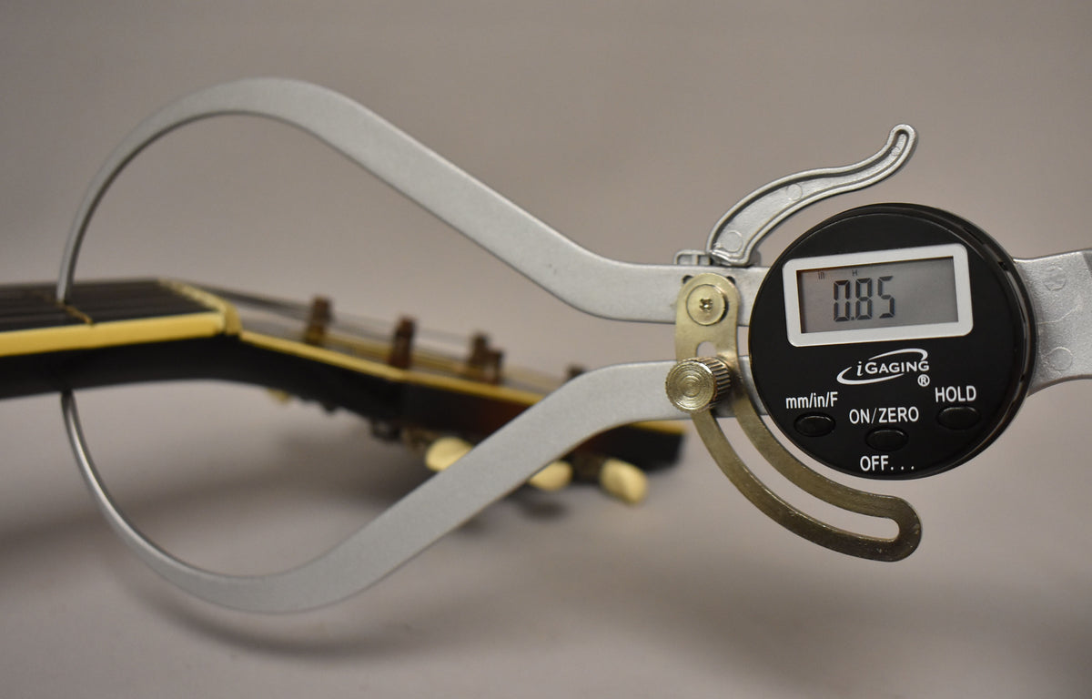 A digital caliper measures the guitar's nut width at 0.85, with the headstock and tuning pegs blurred in the background.