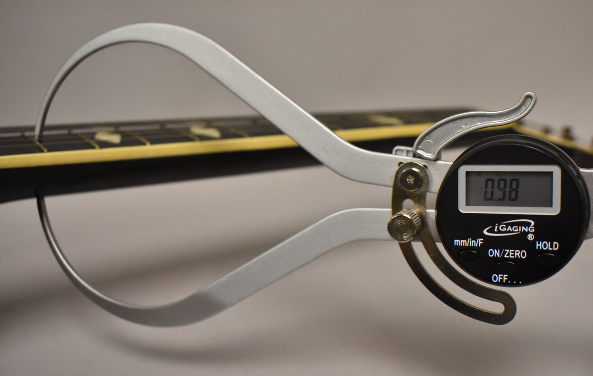 A digital caliper measures the neck thickness at the 1st fret, showing 0.98 inches, with the dark fretboard and block inlays visible.