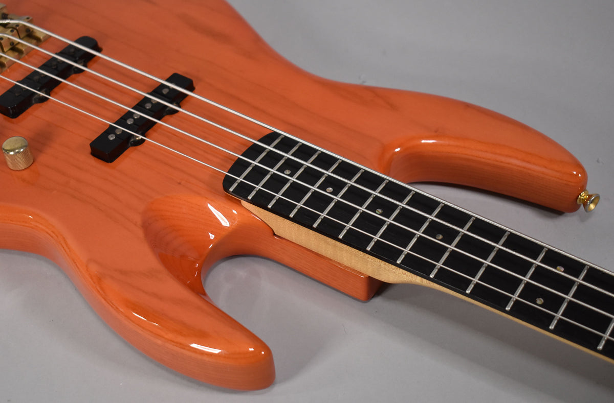 Close-up of the translucent orange bass body, showing the neck joint, two black pickups, a gold control knob, and the lower frets.