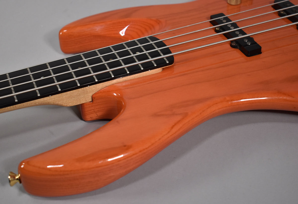 Close-up of the translucent orange bass body and neck joint, showing wood grain, black pickups, and a gold strap button.