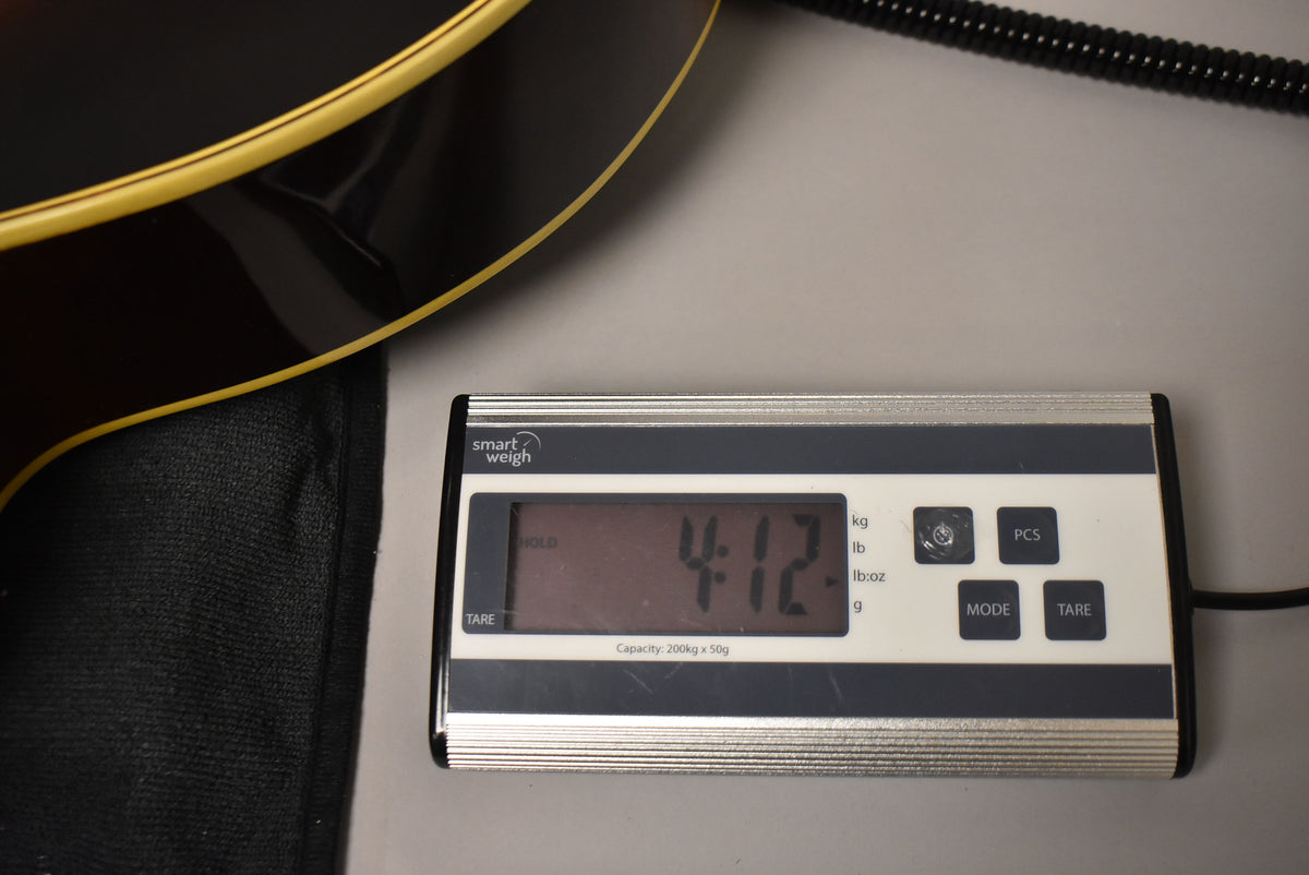 A close-up of the guitar's dark brown body edge with cream binding, resting on a digital scale displaying "4.12" kg.