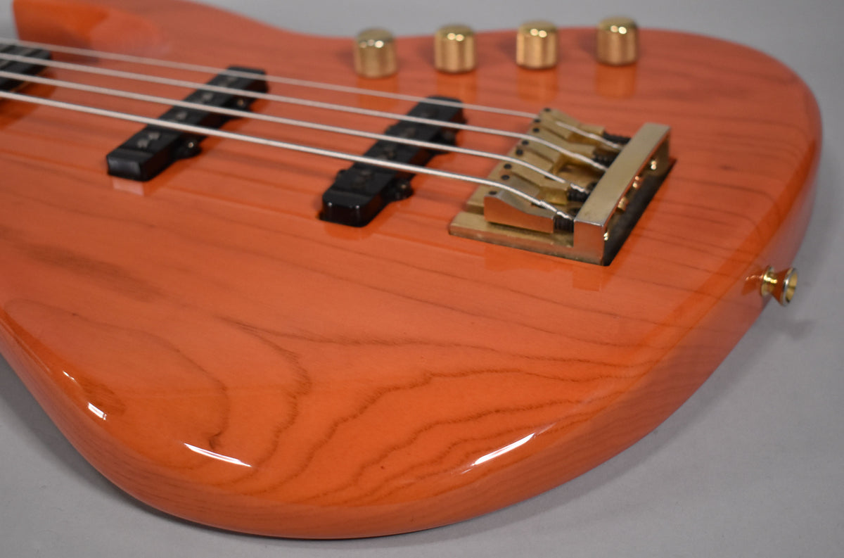 Close-up of the translucent orange bass body, showing wood grain, gold hardware, two black pickups, and four gold control knobs.