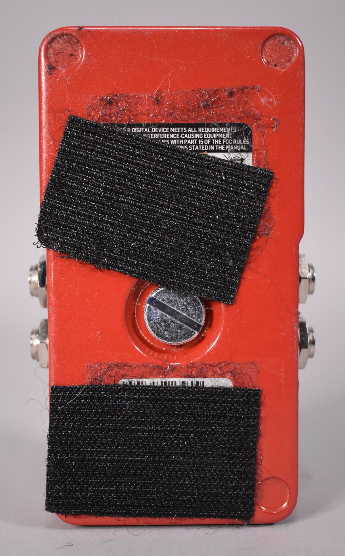The pedal's red underside, covered in two strips of black hook-and-loop fastener, shows a central screw and two silver input jacks.