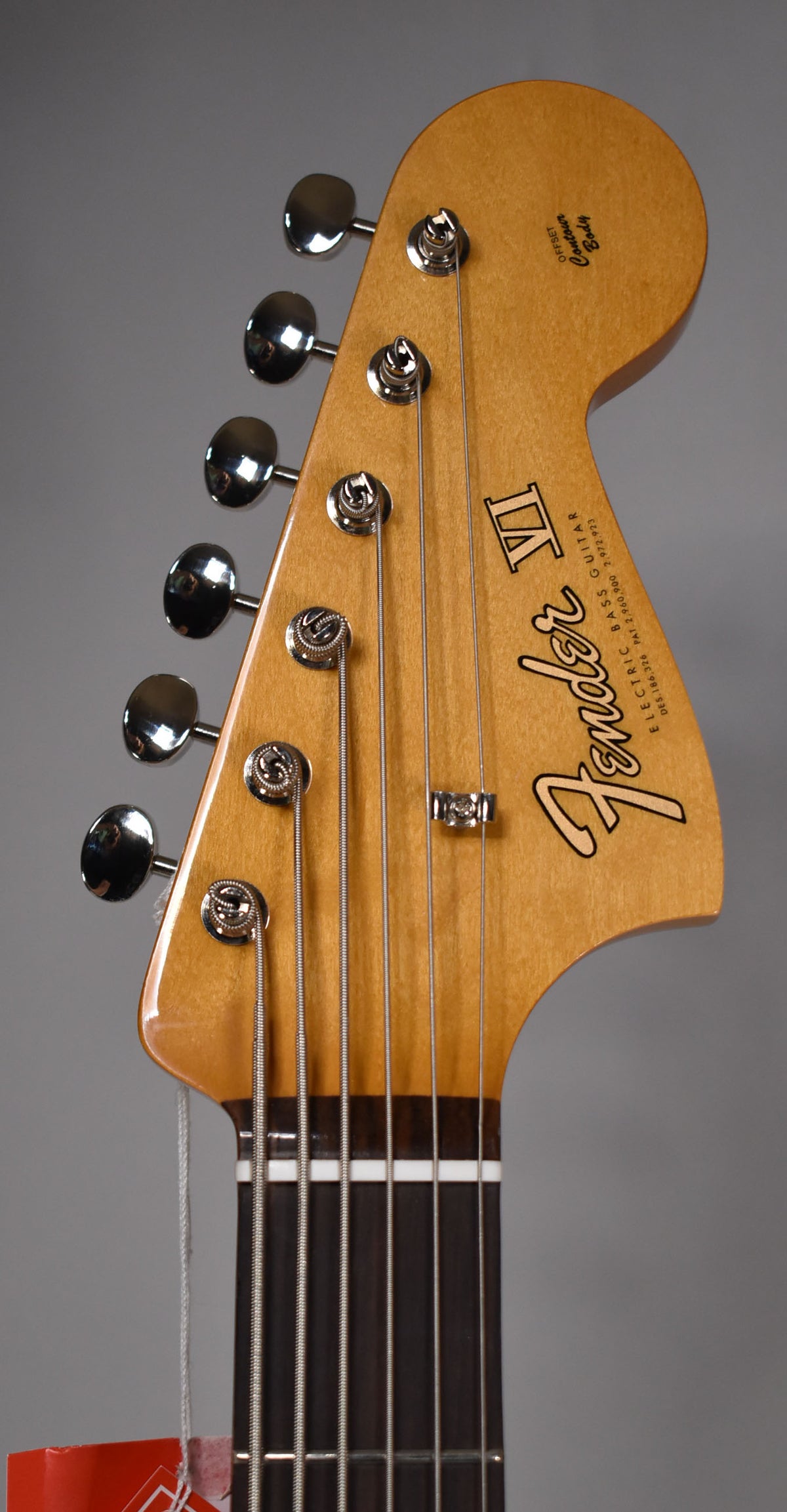 Close-up of the bass guitar's headstock, angled slightly right, showing six chrome tuners, string tree, and a dark rosewood fretboard.