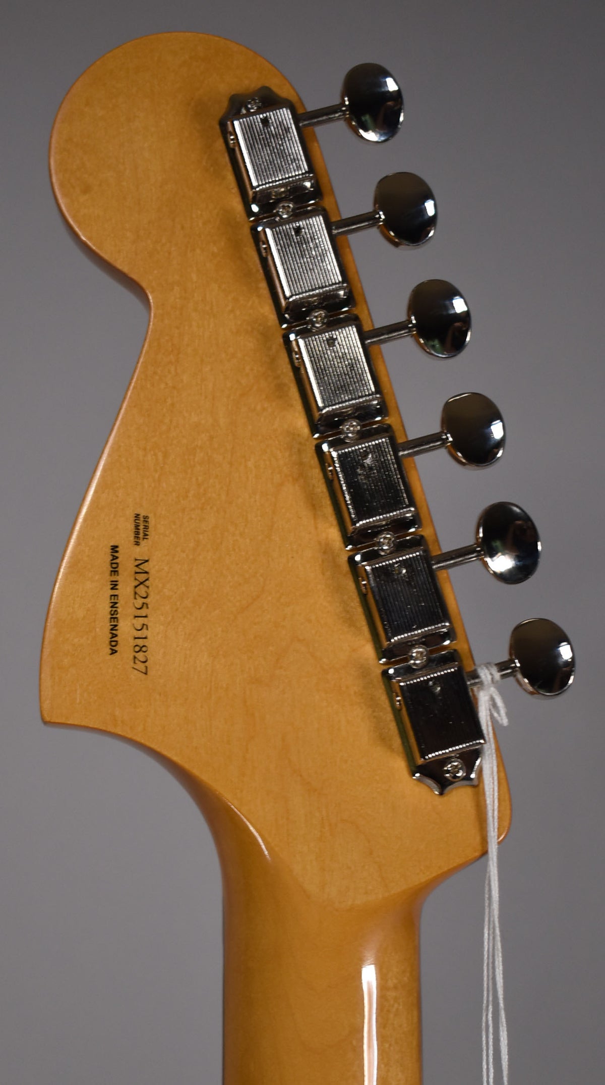 The back of a light brown wooden headstock with six chrome tuners and a white string tied around the lowest tuner.