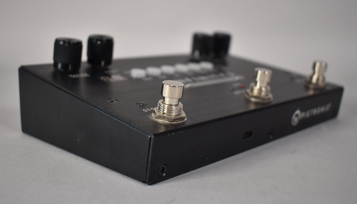 The black brushed metal pedal is angled from the left, showing three silver footswitches and four black control knobs.