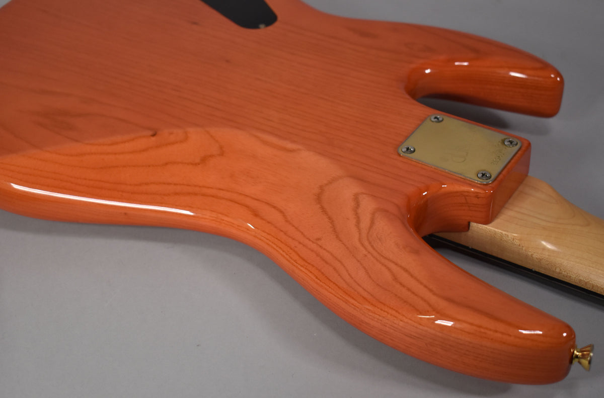 The translucent orange bass body, showing wood grain, has a glossy finish, a brass neck plate, and a gold strap button.