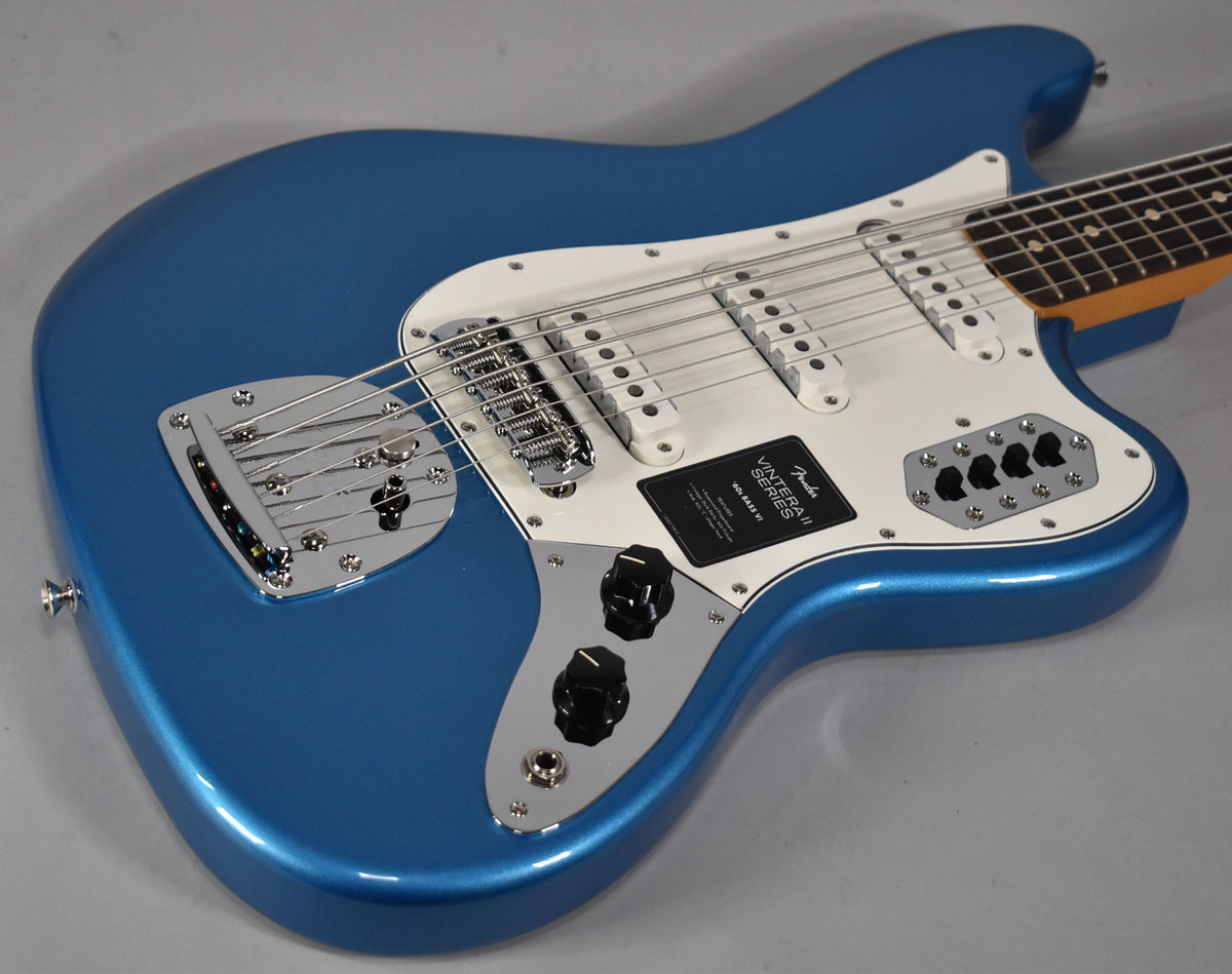 The Lake Placid Blue Bass VI is angled, showing its glossy body, white pickguard, three single-coil pickups, and chrome bridge.