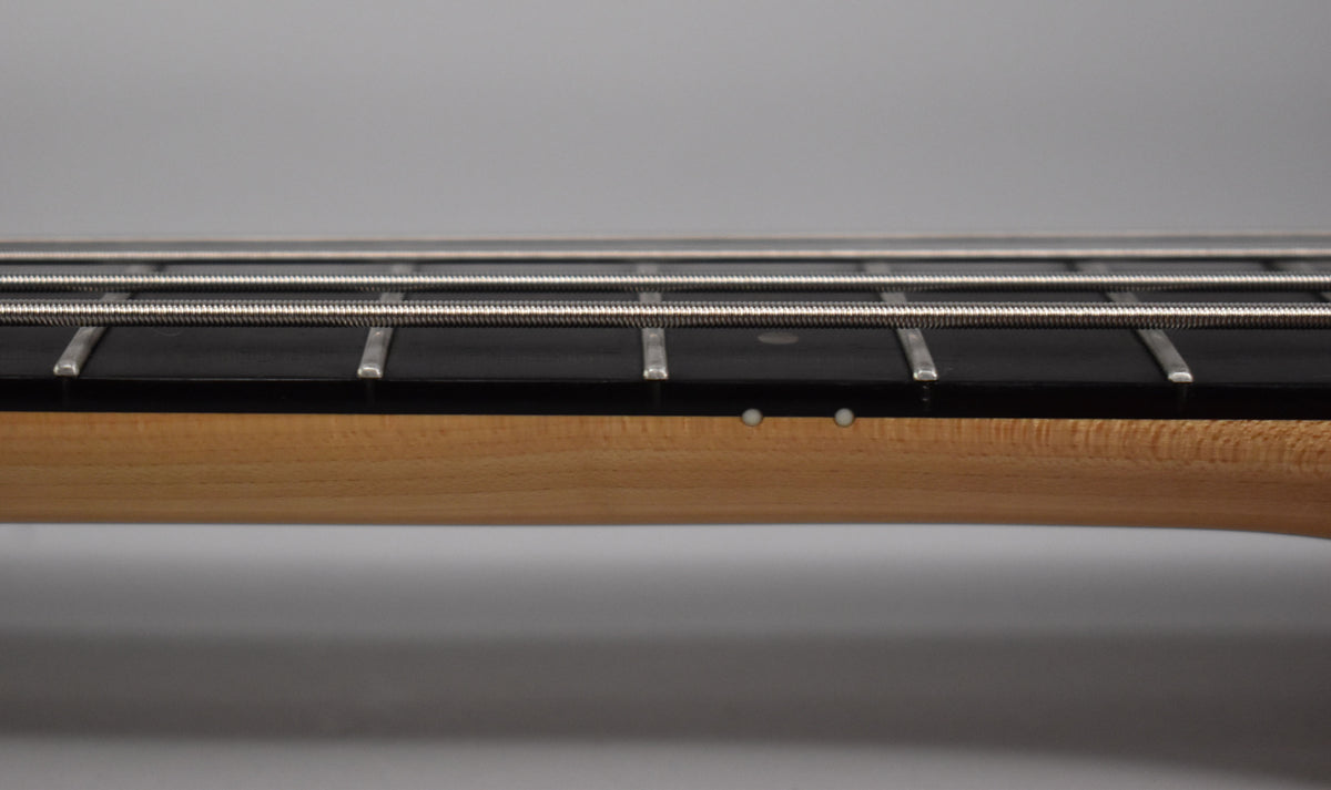 Close-up of the bass neck from the side, showing the light wood neck, dark fretboard, frets, strings, and two white side dots.