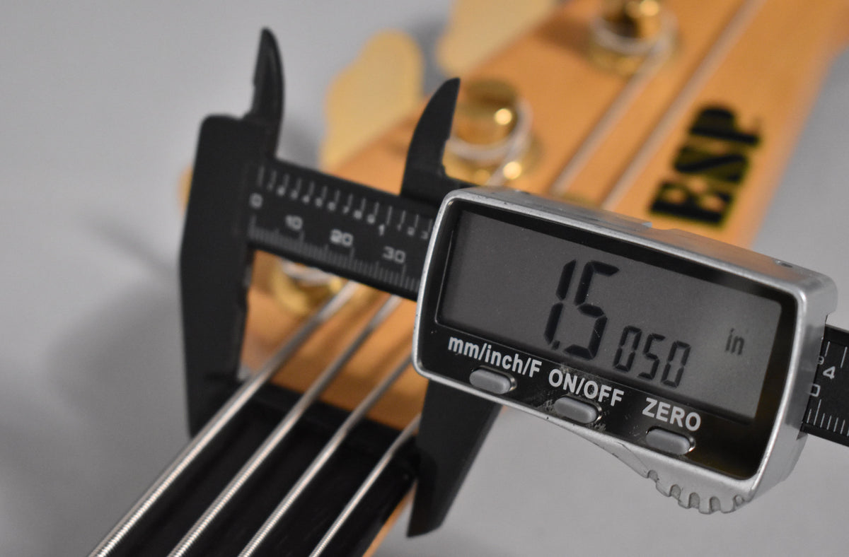 A digital caliper measures the bass's nut width at 1.5050 inches, with the headstock and tuning pegs visible in the background.