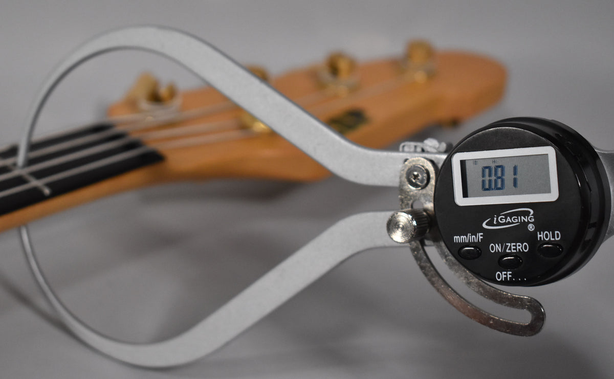 A digital caliper measures the bass's neck relief, showing 0.81mm, with the headstock and tuners blurred in the background.