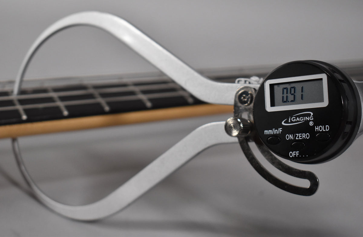 A digital caliper measures the bass's fret height, displaying "0.91" on its screen, with the fretboard and frets visible.