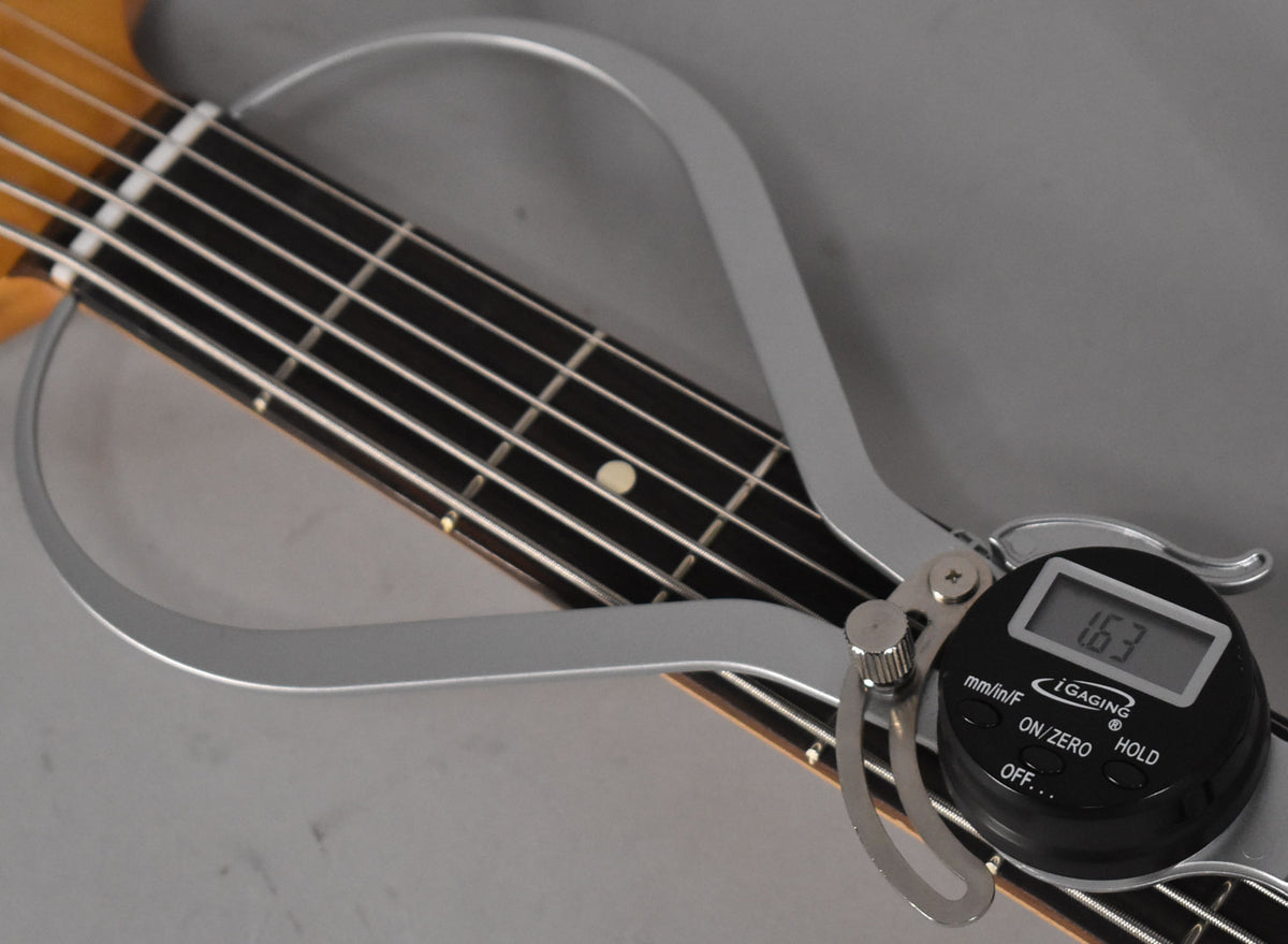 A digital caliper measures the nut width of the bass guitar's dark fretboard, showing "1.63" on its display.