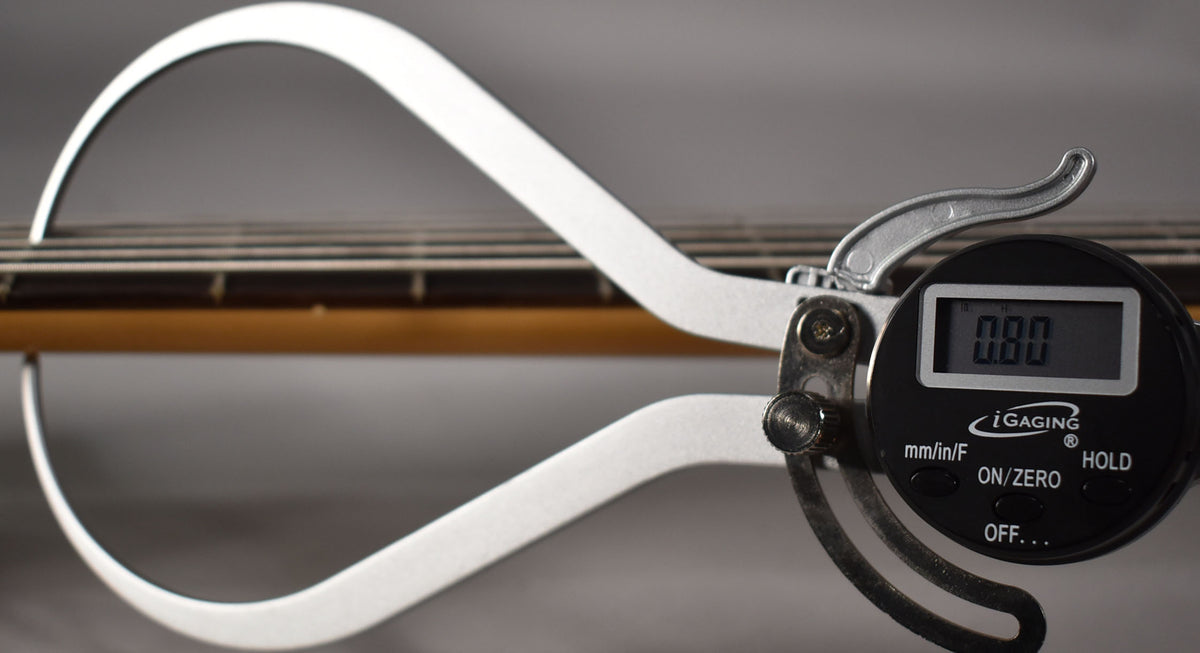 A close-up of a digital caliper measuring the string height on the bass guitar's neck, showing a reading of 0.80.
