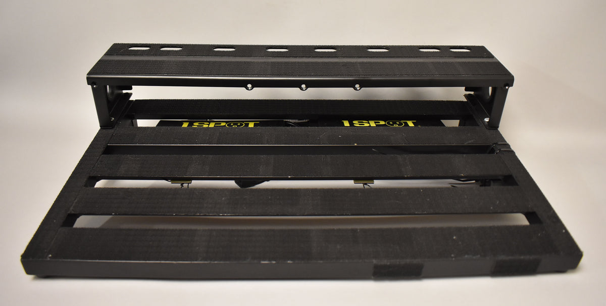 A black pedalboard, angled slightly from the front, showing its two tiers and the yellow "1 SPOT" power supply mounted underneath.