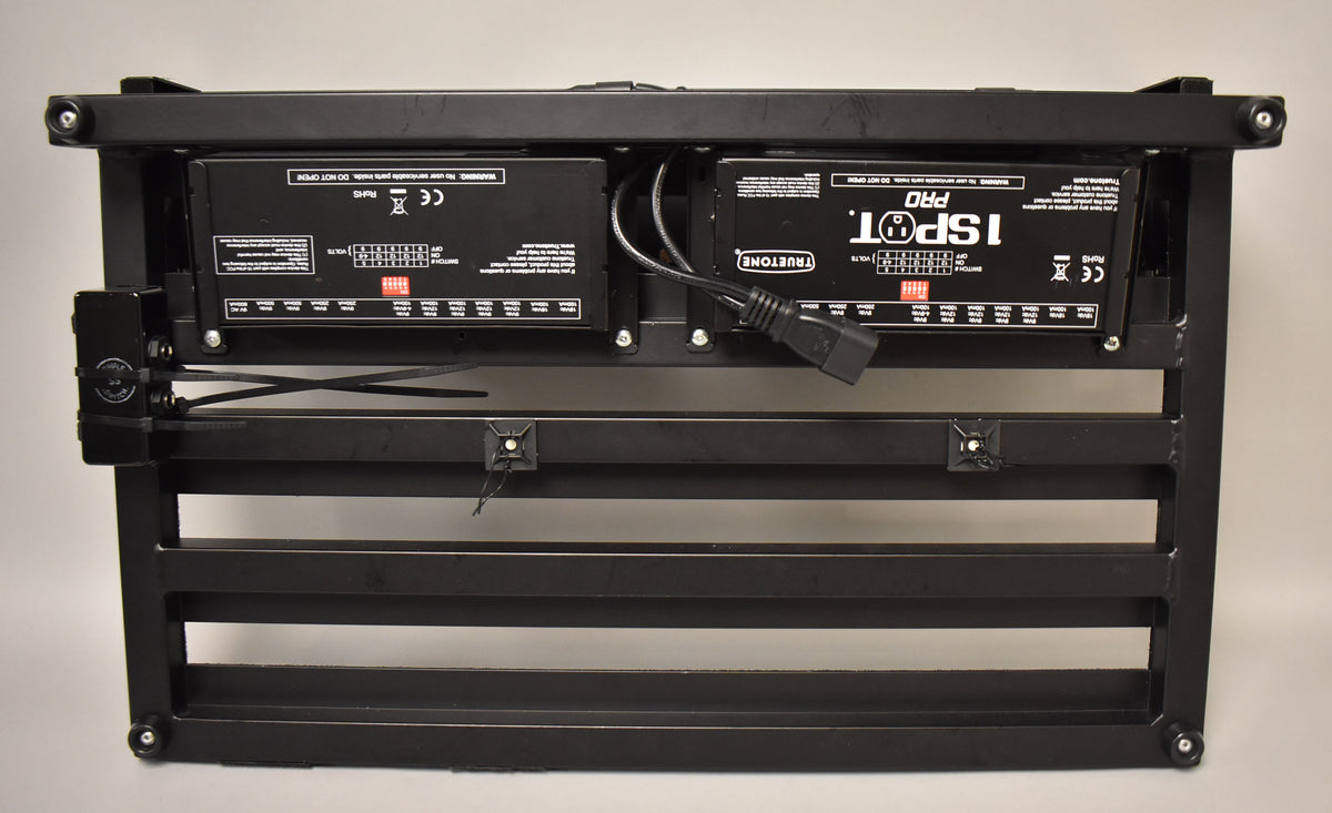 The black pedalboard is seen from below, revealing two power supply units mounted underneath with cables.