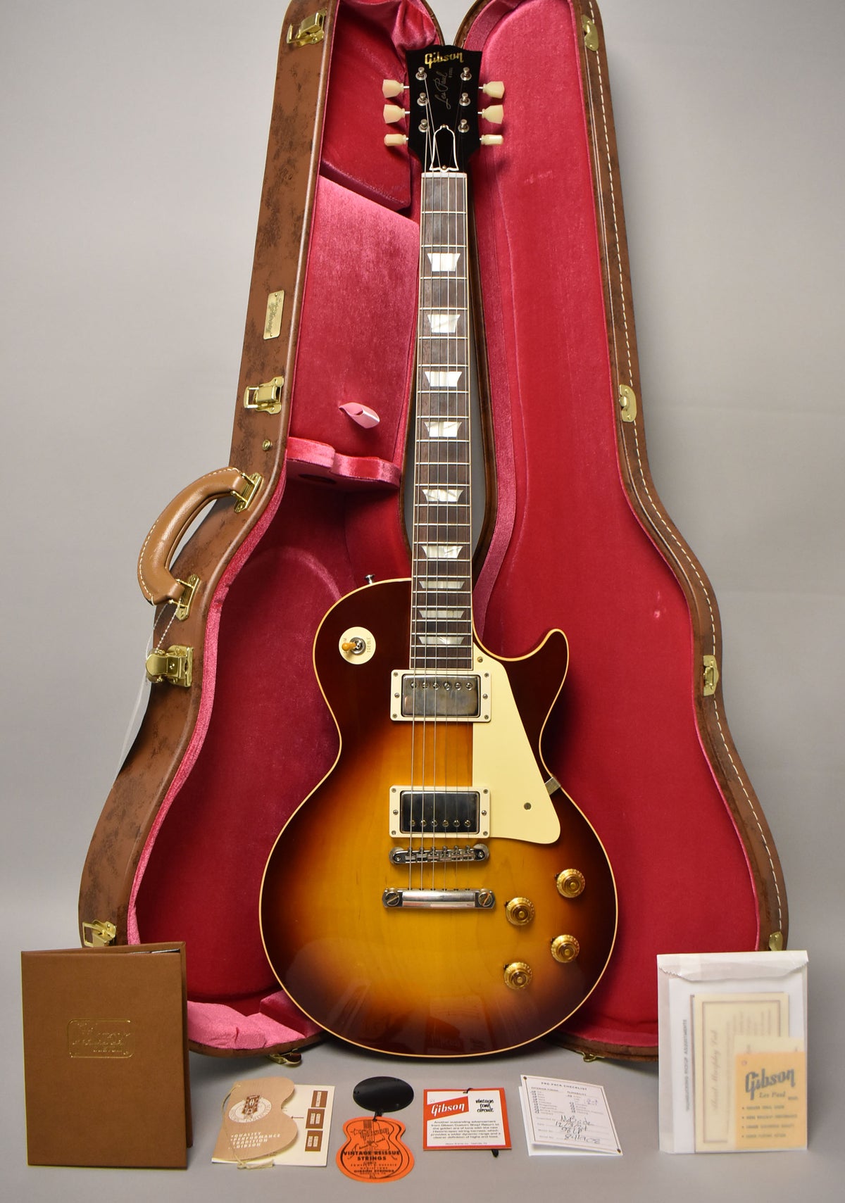 Full-length view of a sunburst electric guitar with cream binding and hardware, resting in a red velvet-lined brown hard case.