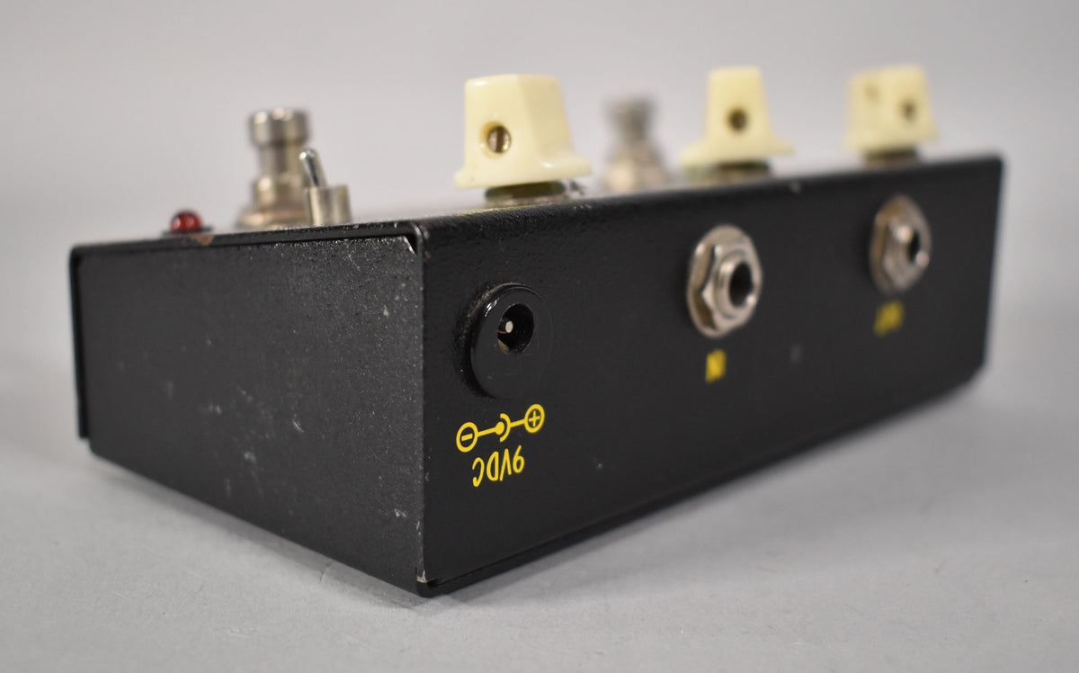 The black textured pedal is angled to show its side, revealing a power input, two audio jacks, and yellow 9VDC lettering.