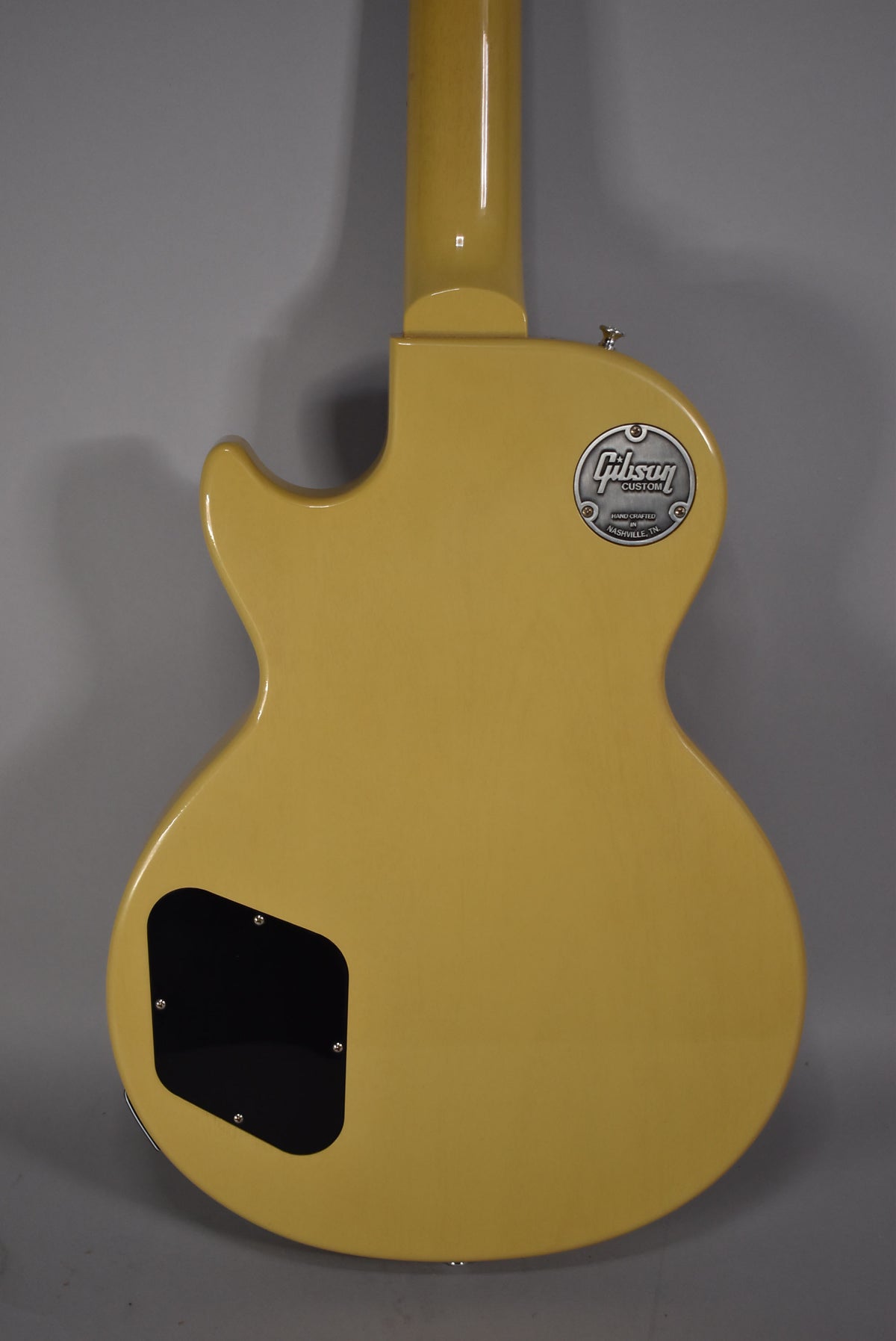 The TV yellow guitar's back is shown, with a black control cavity cover and a silver Gibson Custom medallion near the neck joint.