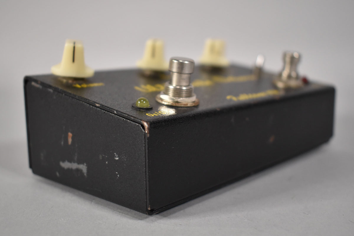 Close-up of the black pedal's right side, showing a green LED, silver footswitch, and three cream knobs, with paint chips.