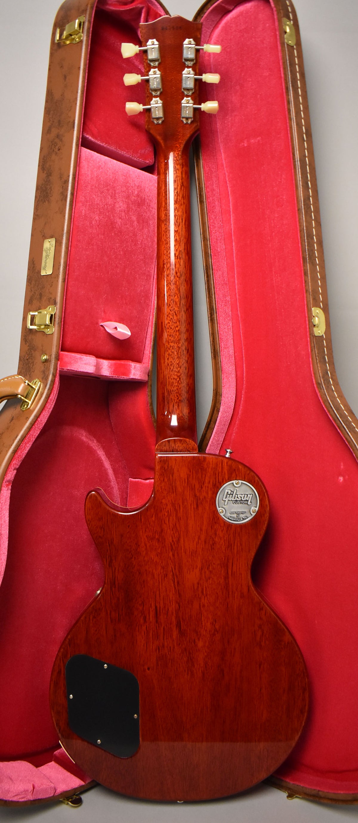 The back of a glossy, dark reddish-brown guitar with a black control cavity cover, resting in a brown case with red velvet lining.