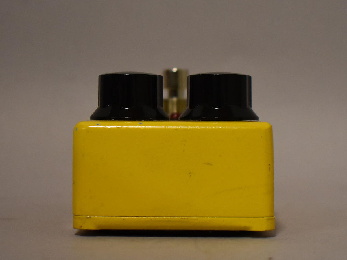 A yellow guitar pedal with two black knobs on top, viewed from the side against a gray background.
