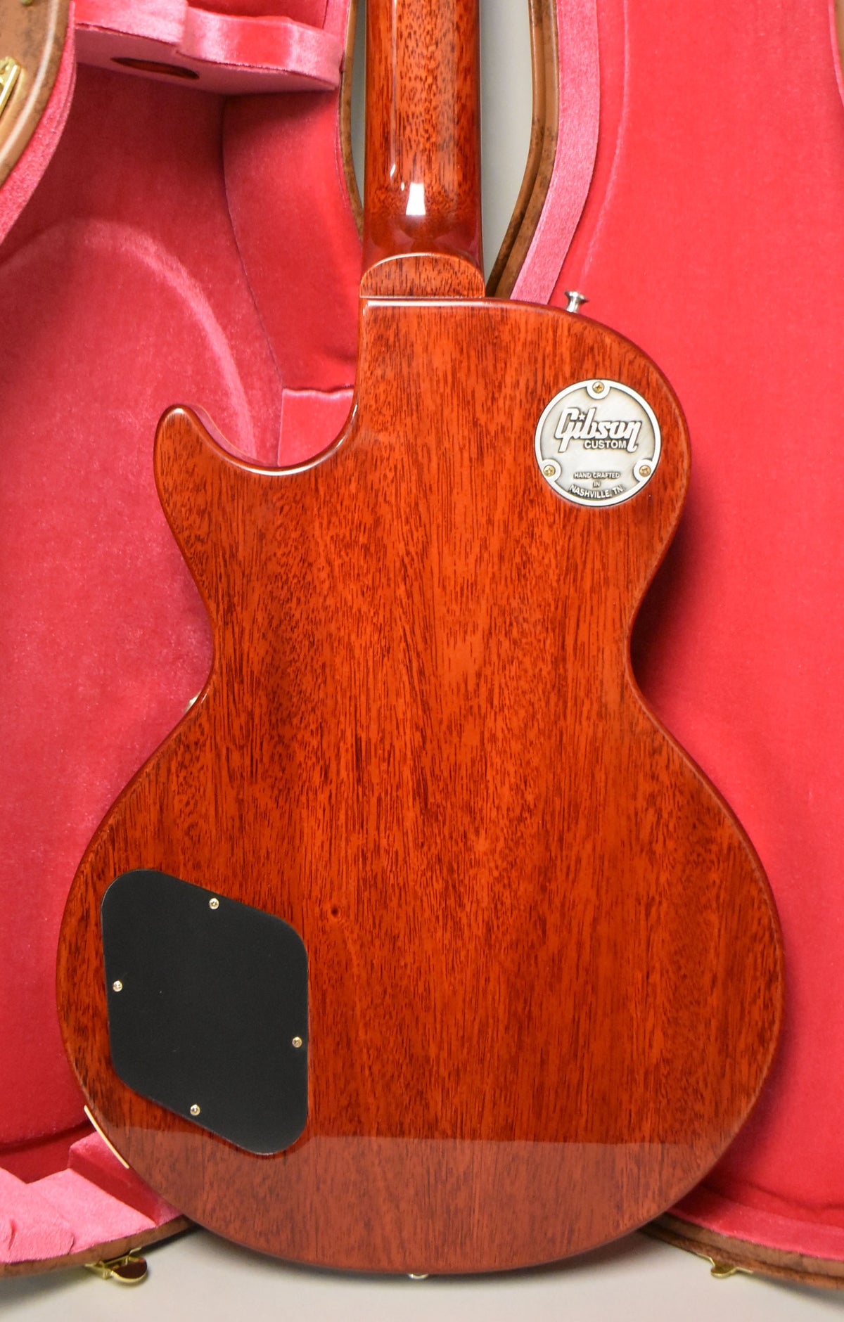 The guitar's back, a rich bourbon burst with visible wood grain, is framed in a red velvet-lined case, showing a black control plate.