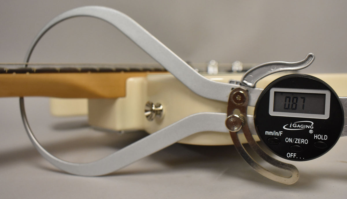 A digital caliper measures the guitar's neck thickness at 0.87 inches, with the blonde body and maple neck visible.
