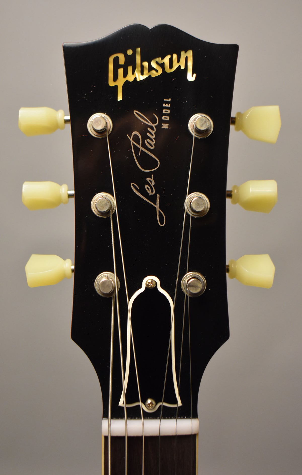 A close-up of the black headstock, showing six cream tuning pegs, a gold bell-shaped truss rod cover, and light finish checking.