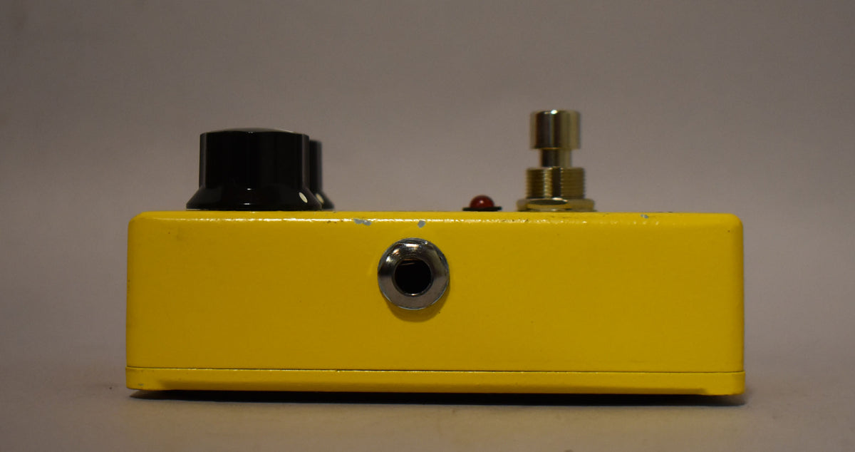 The yellow pedal is viewed from the side, showing a black knob, a red LED, a gold footswitch, and a silver input jack.