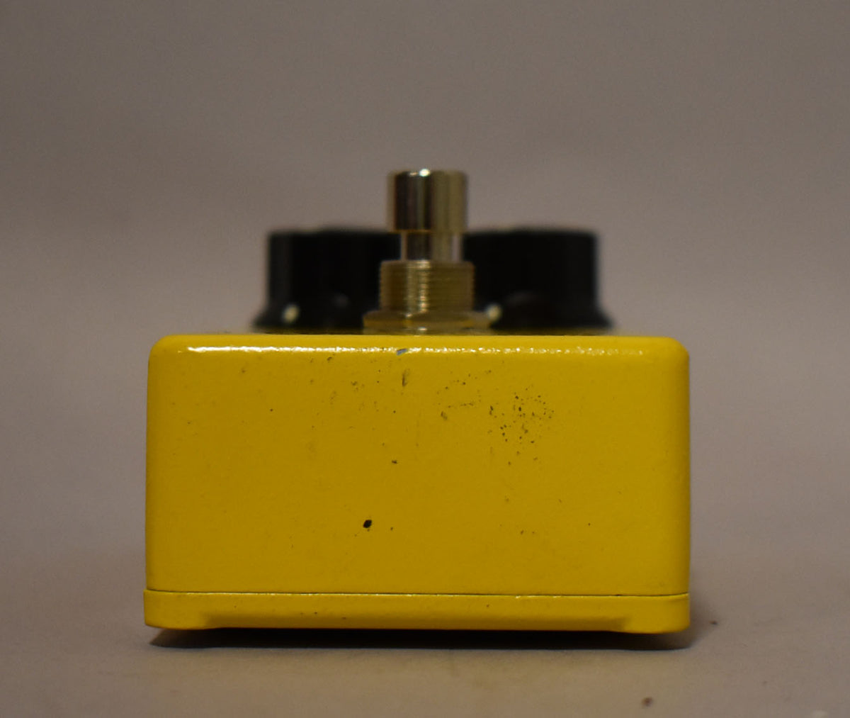 The yellow pedal is viewed from the side, showing a gold knob shaft and black knobs blurred in the background, with minor scuffs.