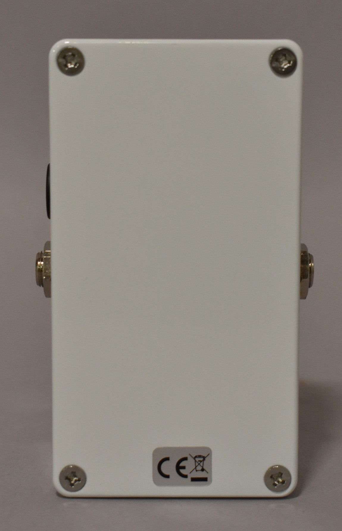 The white effect pedal is viewed from the back, showing four silver screws, two input jacks, and a CE mark sticker.