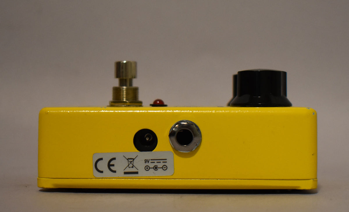 The bright yellow pedal is viewed from the back, showing a power input, a jack, and a 9V sticker.