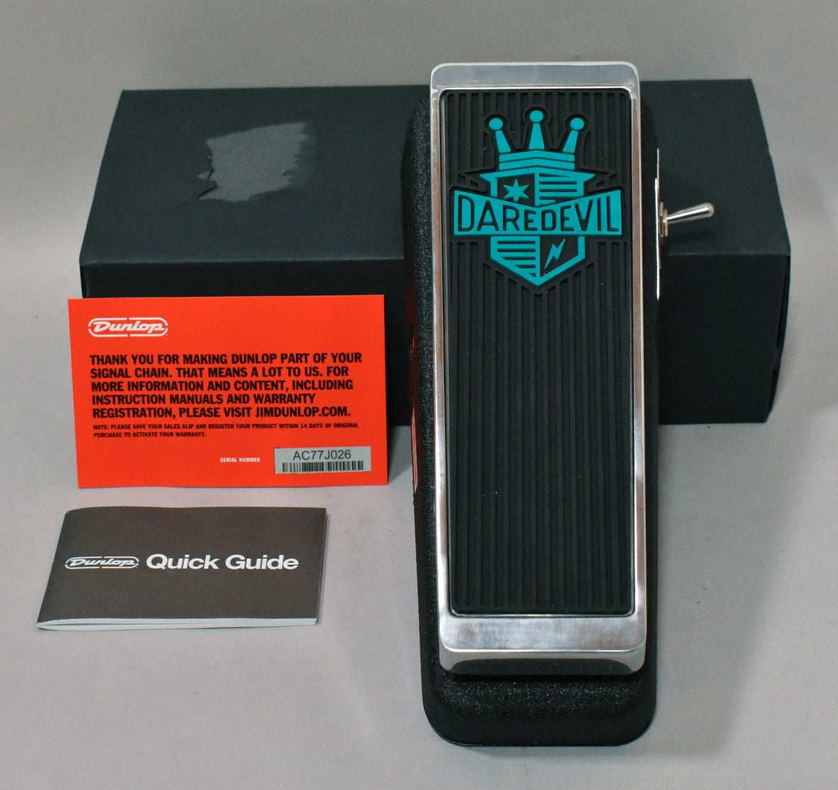 A black wah pedal with a ribbed rubber treadle, chrome trim, and a teal "Daredevil" logo, shown with its box and paperwork.
