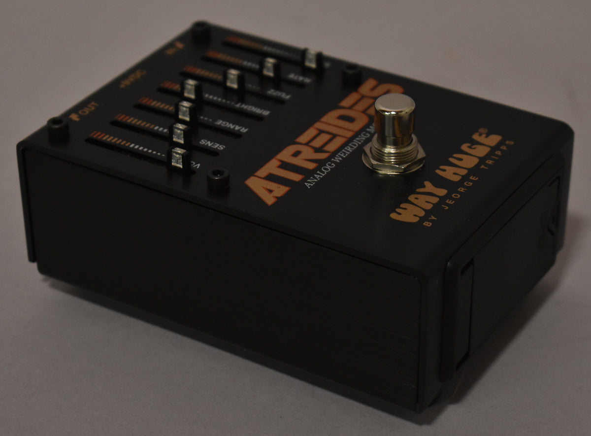 A black guitar pedal is angled from the right, showing its top with five horizontal fader controls and a silver footswitch.