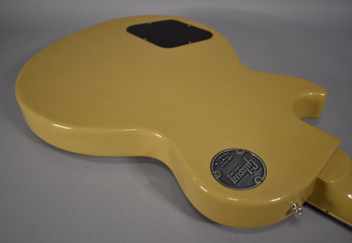 The TV yellow guitar's back is angled, showing a black control cavity cover and a silver Gibson Custom medallion near the strap button.