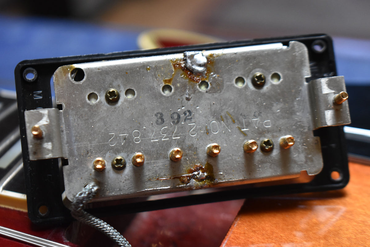 Close-up of the underside of a humbucker pickup, showing the metal baseplate with visible solder joints and some corrosion.