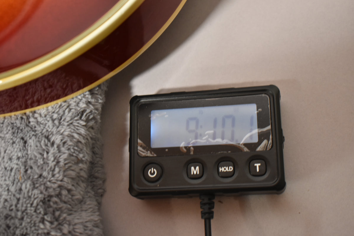 A digital scale displays "9.10" next to the cherry sunburst body binding of the guitar, resting on a gray plush surface.