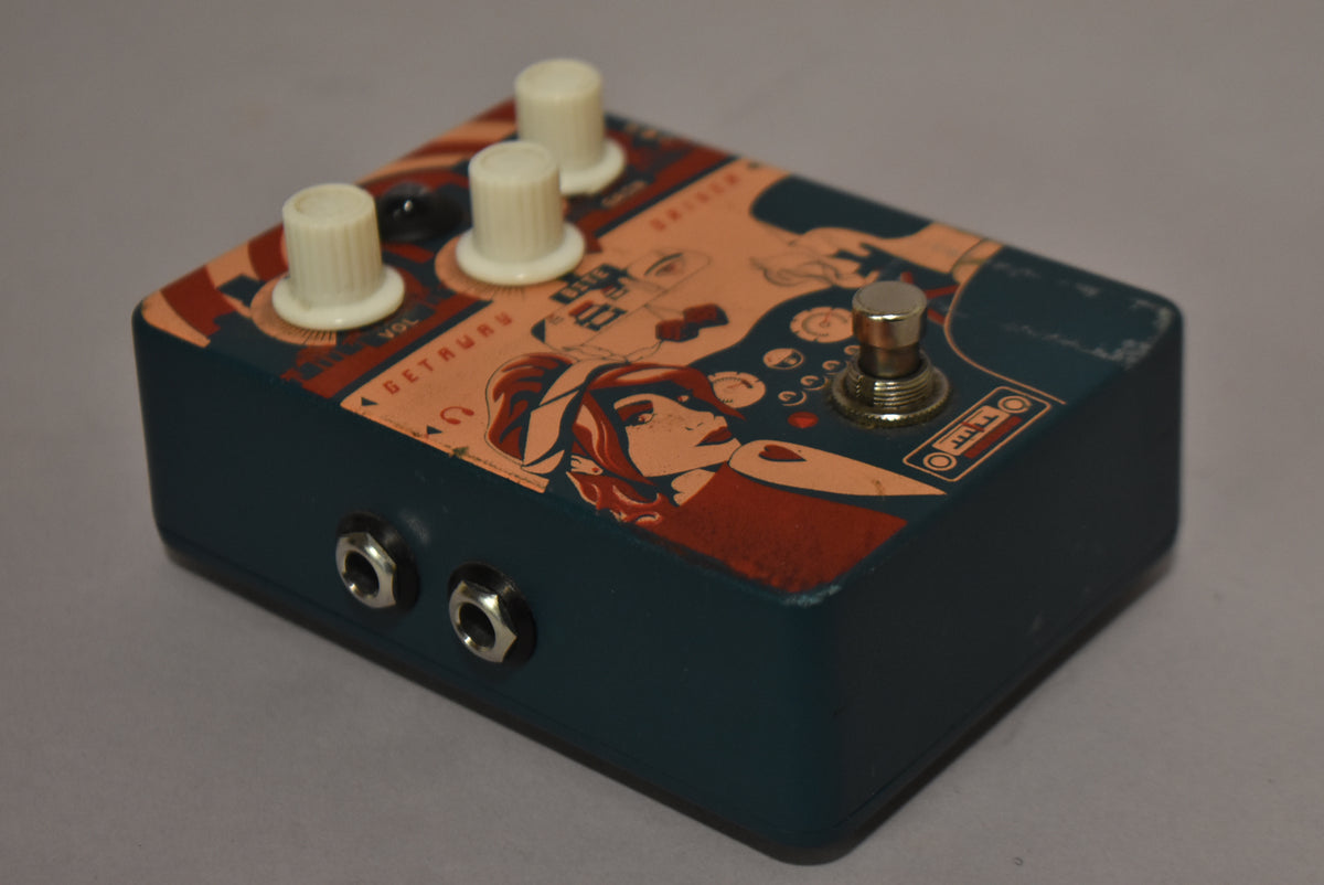 The pedal is angled from the left, showing its dark teal sides and top with a retro graphic, three cream knobs, and a footswitch.