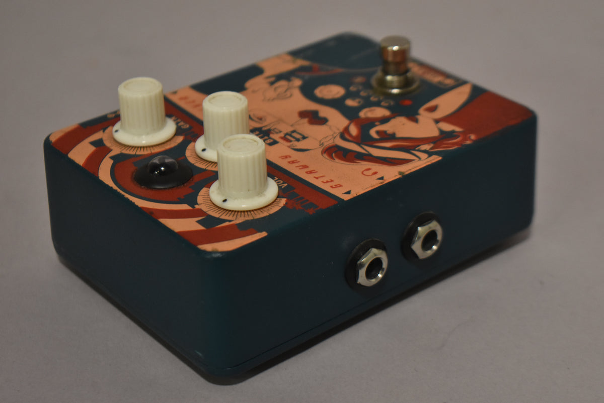 The dark teal pedal is angled right, showing the top with three cream knobs, a black LED, and a footswitch, plus two input jacks on the right side.