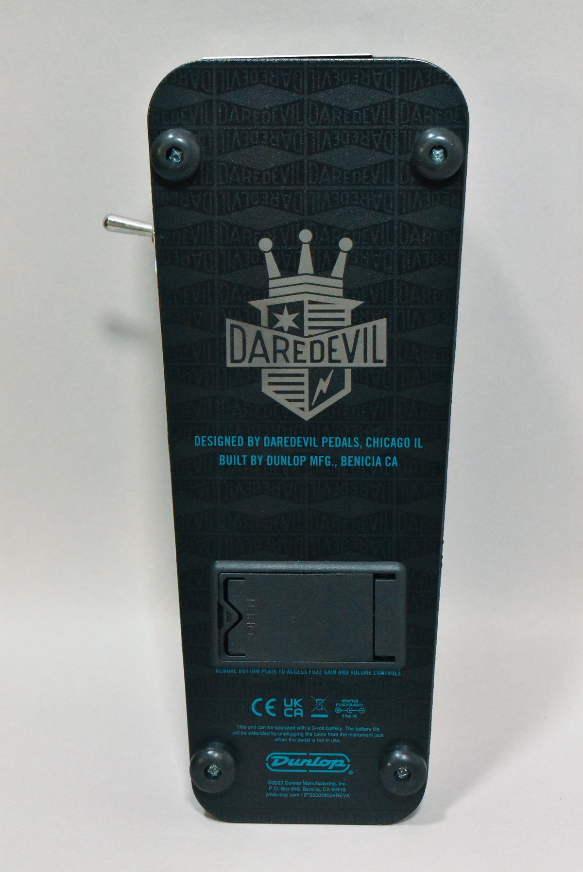 The pedal's black textured underside is shown, with a battery compartment, four rubber feet, and a metal toggle switch on the left.